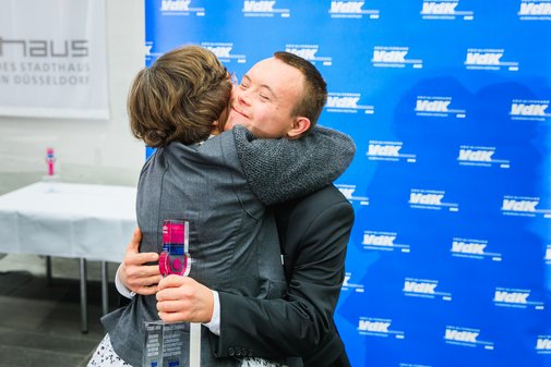 Ein Mann mit vilmA-Pokal in der Hand wird von einer anderen Person innig umarmt. Im Hintergrund die blaue Pressewand des VdK mit Logos. 