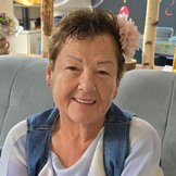 Porträt einer lächelnden älteren Frau mit kurzen braunen Haaren und hellblauem Pullover und Jeansweste. Sie sitzt auf einem hellgrauen Sofa.