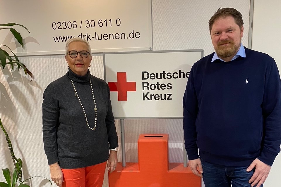 Eine Frau mit kurzen Haaren und dunkler Brille und ein Mann mit kurzem braunen Bart stehen vor einem Schild des Deutschen Roten Kreuzes. Im Hintergrund ist eine Webseite und Telefonnummer des DRK Lünen zu sehen.
