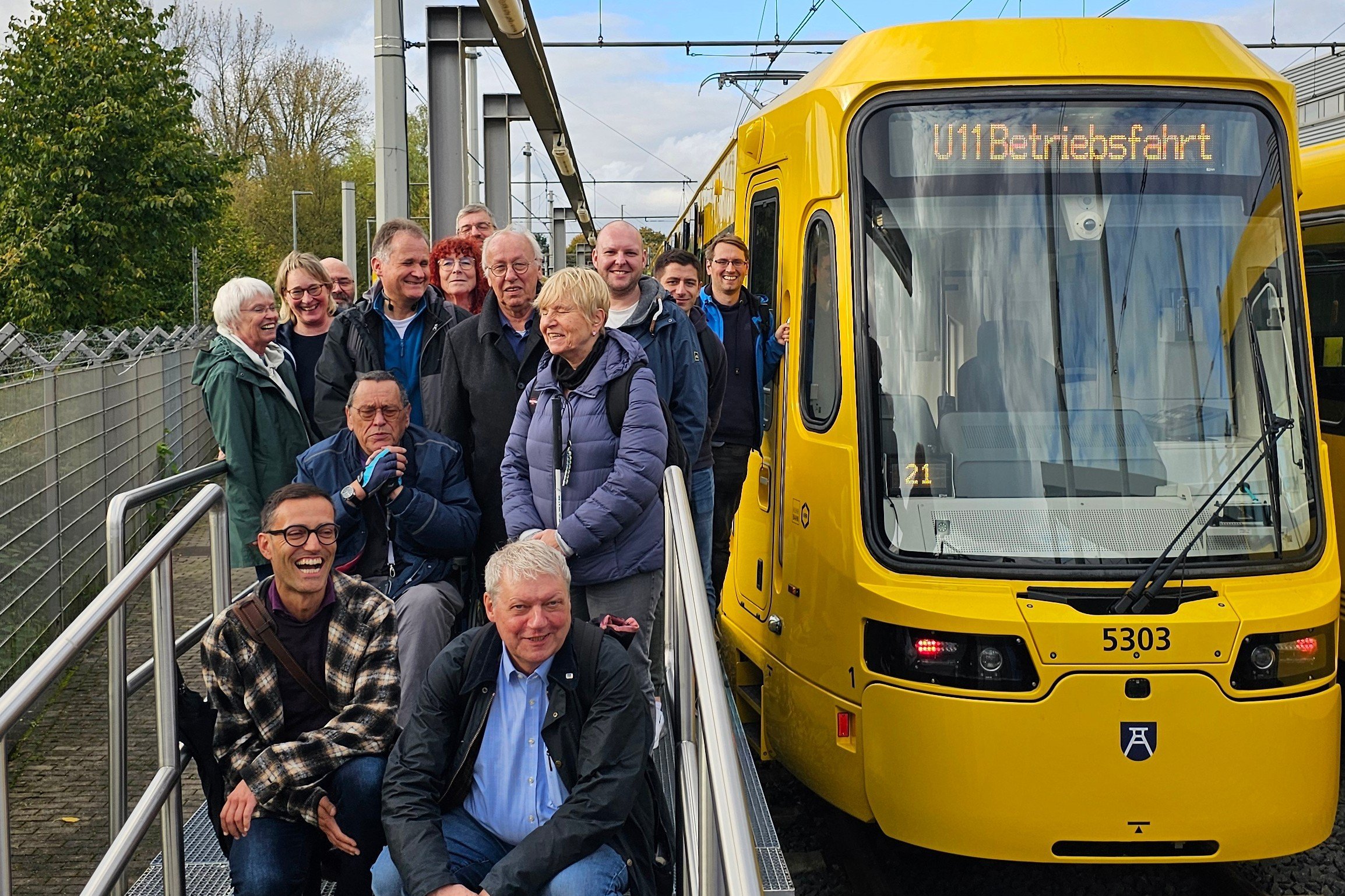 Eine Gruppe Männer und Frauen hockt und steht für ein Gruppenfoto auf der neuen Rollstuhl-Zufahrt neben einem der neuen gelben Ruhrbahn-Fahrzeuge.