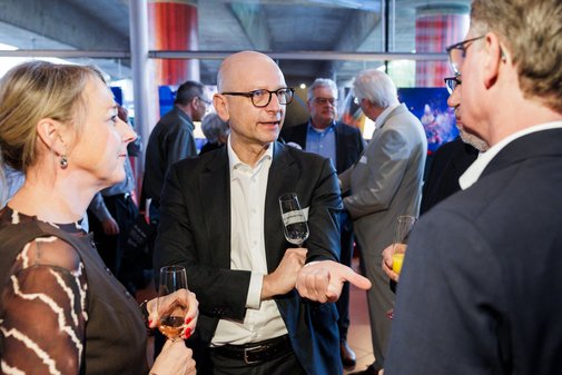 eine Frau mit blond-brauen Haaren l&auml;chelt einen Mann an. In der Mitte steht zudem ein Mann ohne Haare und mit Brille.