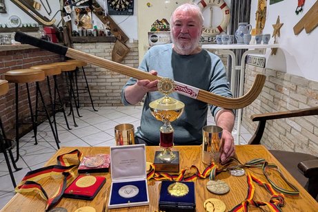 Werner Achtig mit Hockeyschl&auml;ger in der Hand und Medaillen vor sich auf dem Tisch