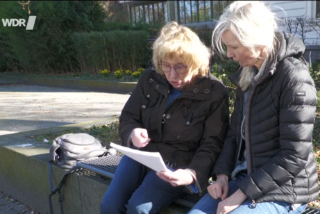Zwei Frauen sitzen nebeneinander auf einer Betonmauer und schauen auf ein Blatt Papier