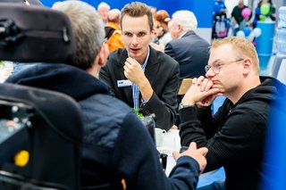 Drei Menschen, davon einer im Rollstuhl, sitzen im Gespräch an einem Tisch des VdK-Stands.