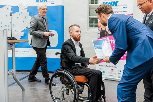 Einer der Laudatoren überreicht einem Preisträger im Rollstuhl eine Urkunde und schüttelt ihm die Hand.