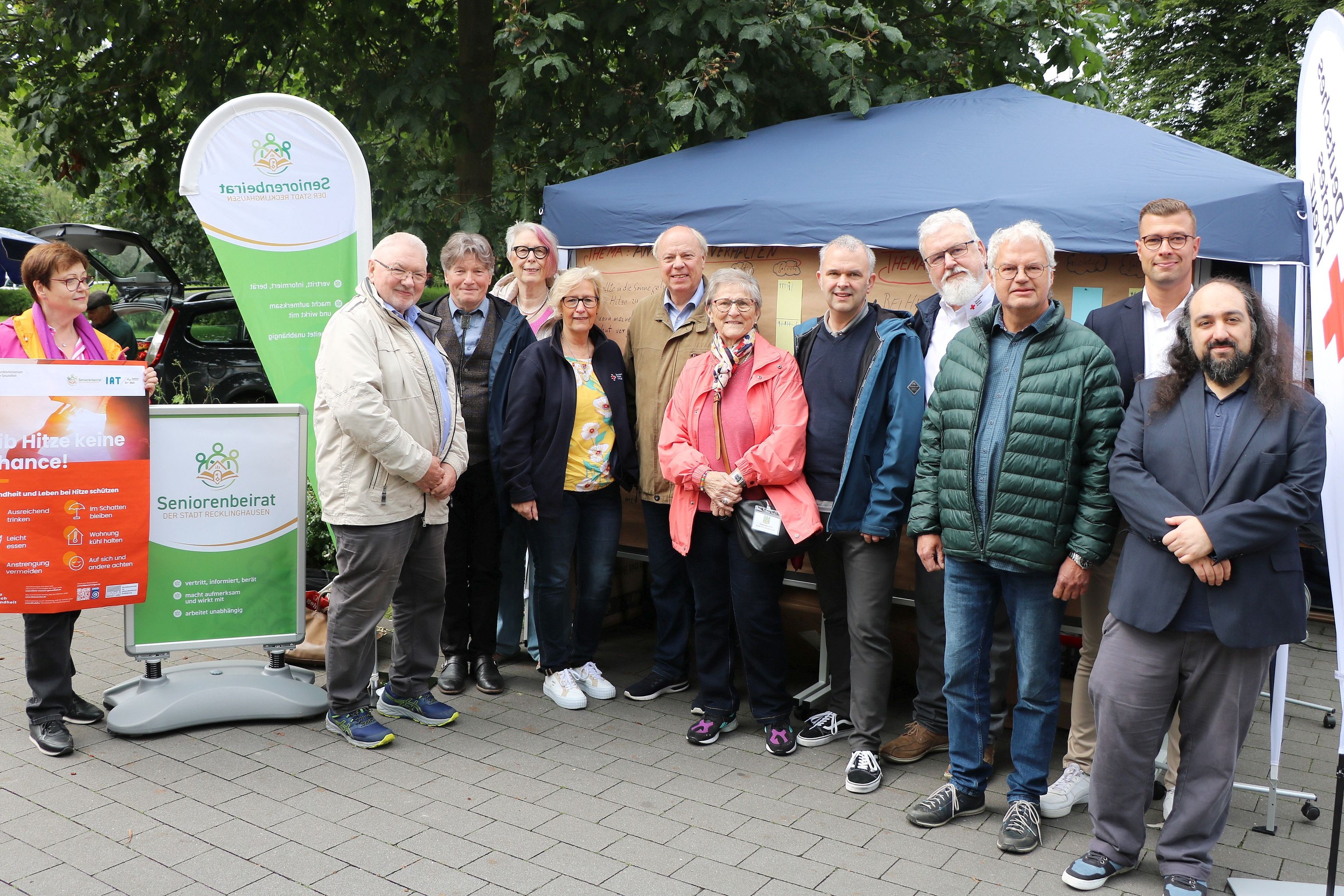 Eine Gruppe Menschen steht vor einem Informationsstand. Eine Dame links im Bild hält ein Plakat zum Thema Hitzeschutz in die Höhe.