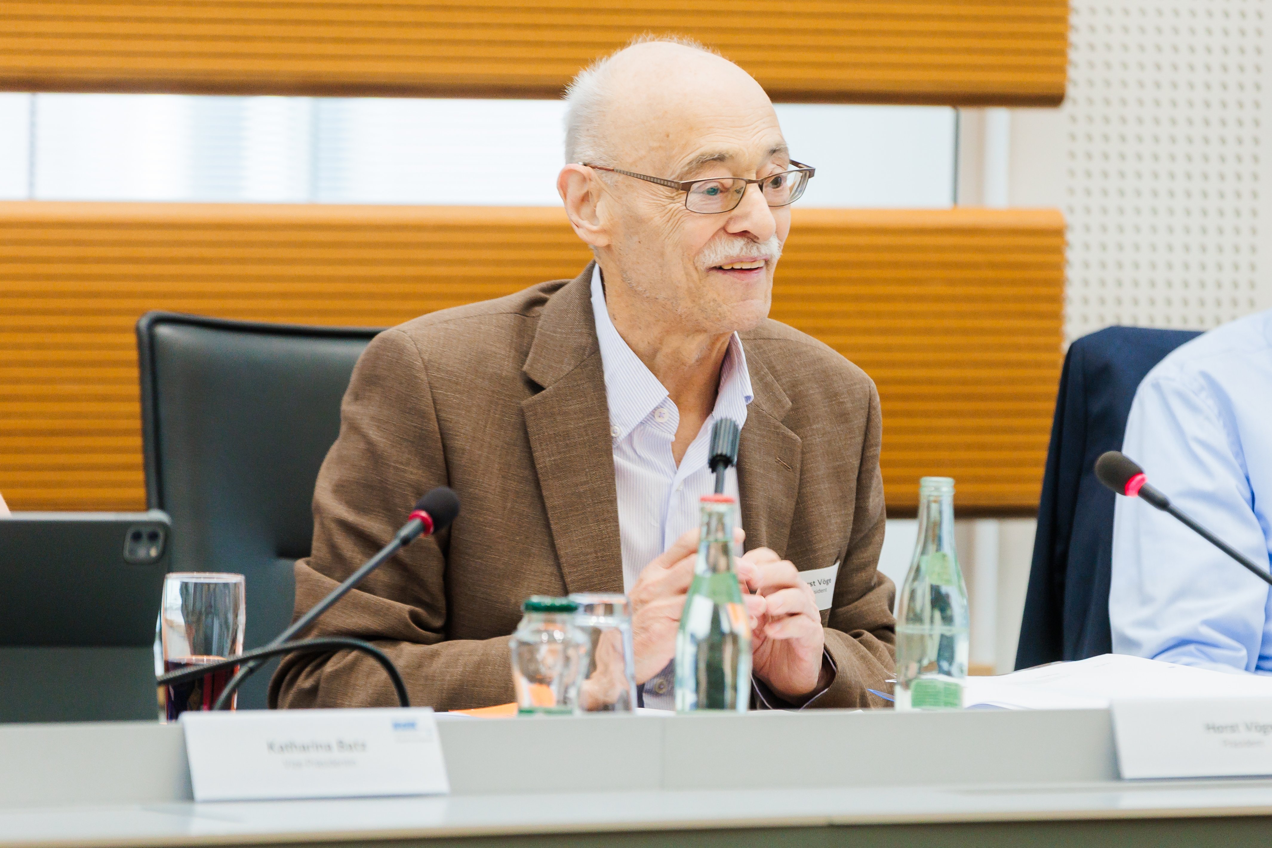 Horst Vöge, ein älterer Herr mit Halbglatze und Brille, sitzt auf dem Podium am Rednerpult und spricht lächelnd Richtung Plenum.