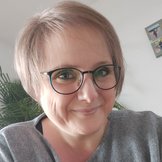 Porträt einer lächelnden Frau mit kurzen blonden Haaren und runder Brille in grauem Oberteil.
