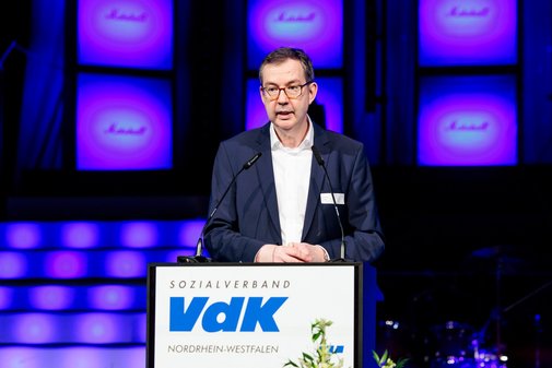 VdK-Vorstandssprecher Thomas Zander steht an einem Pult mit der Aufschrift VdK. Er tr&auml;gt eine Brille, ein Sakko und ein wei&szlig;es Hemd.