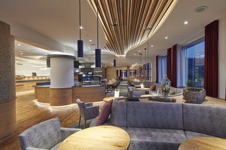 Blick von der mit Sesseln und Sofas ausgestattete Lobby auf einen in Holz und mit vielen Lichtelementen gestalteten Restaurant-Bereich.