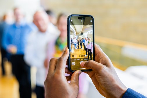 Hände halten ein Smartphone, um ein Foto zu machen. Dahinter verschwommen die Personen, die gerade fotografiert werden. 