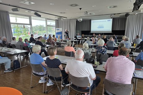 Viele Menschen sitzen an langen Tischen in einem Saal. Vorne ist eine B&uuml;hne mit Leinwand. 