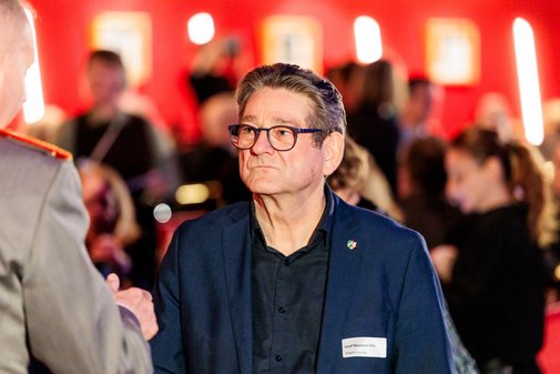 ein Mann mit schwarzen Haaren und Brille spricht mit einem Mann, der links im Bild kaum zu sehen ist.