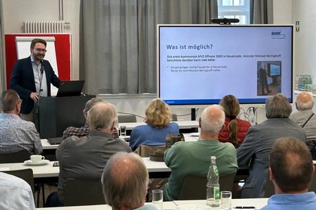 Blick von hinten über die Tischreihen mit Menschen. Vorne im Raum steht ein Referent am Rednerpult, neben sich eine Whiteboard-Präsentation.