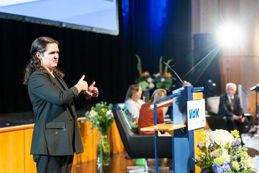 Eine Gebärdendolmetscherin steht am Rand der Bühne und übersetzt die Redebeiträge der Podiumsdiskussionsteilnehmer (im Bildhintergrund).