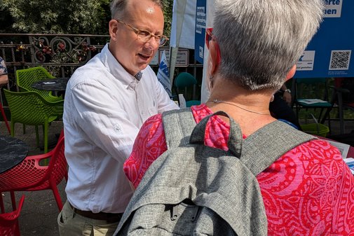 Ein Mann mit Brille und hellem Hemd und eine Frau mit rotem Shirt und grauem Rucksack auf dem Rücken stehen an einem Stehtisch und unterhalten sich.