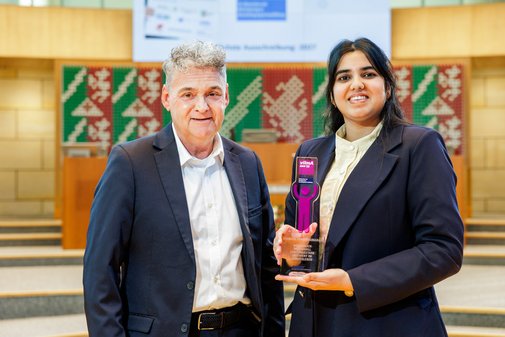 Ein Mann mit dunklem Sakko, Hemd und grauem Kurzhaarschnitte steht neben einer l&auml;chelnden jungen Frau mit dunklen langen Haaren und Sakko, die einen vilmA-Preis in der Hand h&auml;lt.