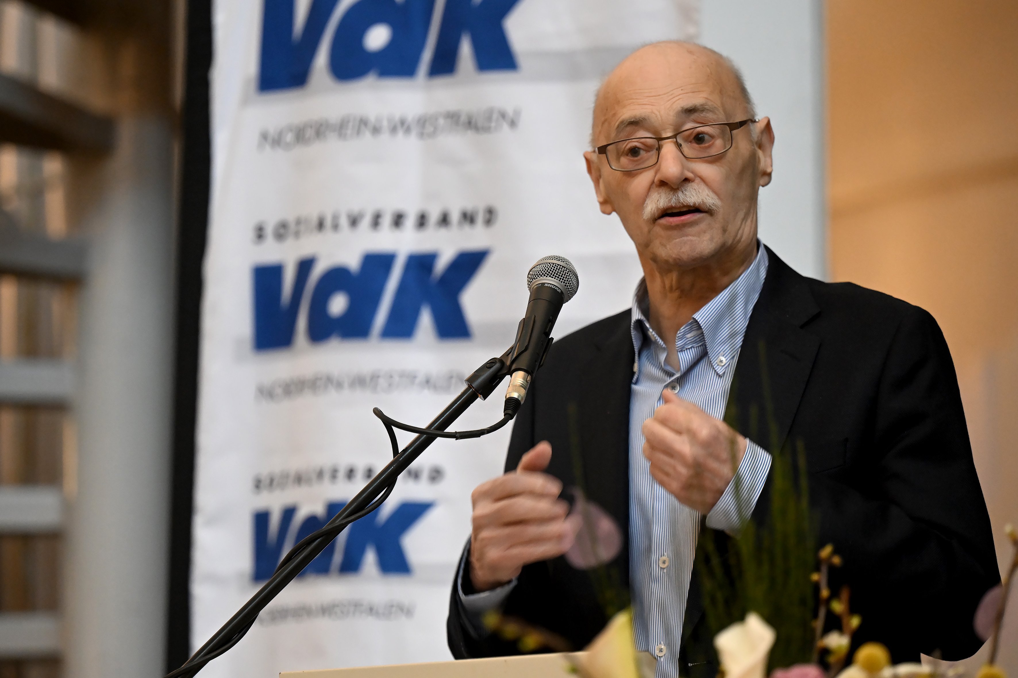 VdK-NRW-Präsident steht am Rednerpult und spricht. Im Hintergrund hängt ein Banner mit VdK-NRW-Logos.