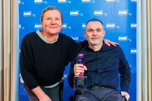 Ein vilmA-Gewinner gemeinsam mit einer Begleitperson vo der blauen VdK-Pressewand mit Logos. 