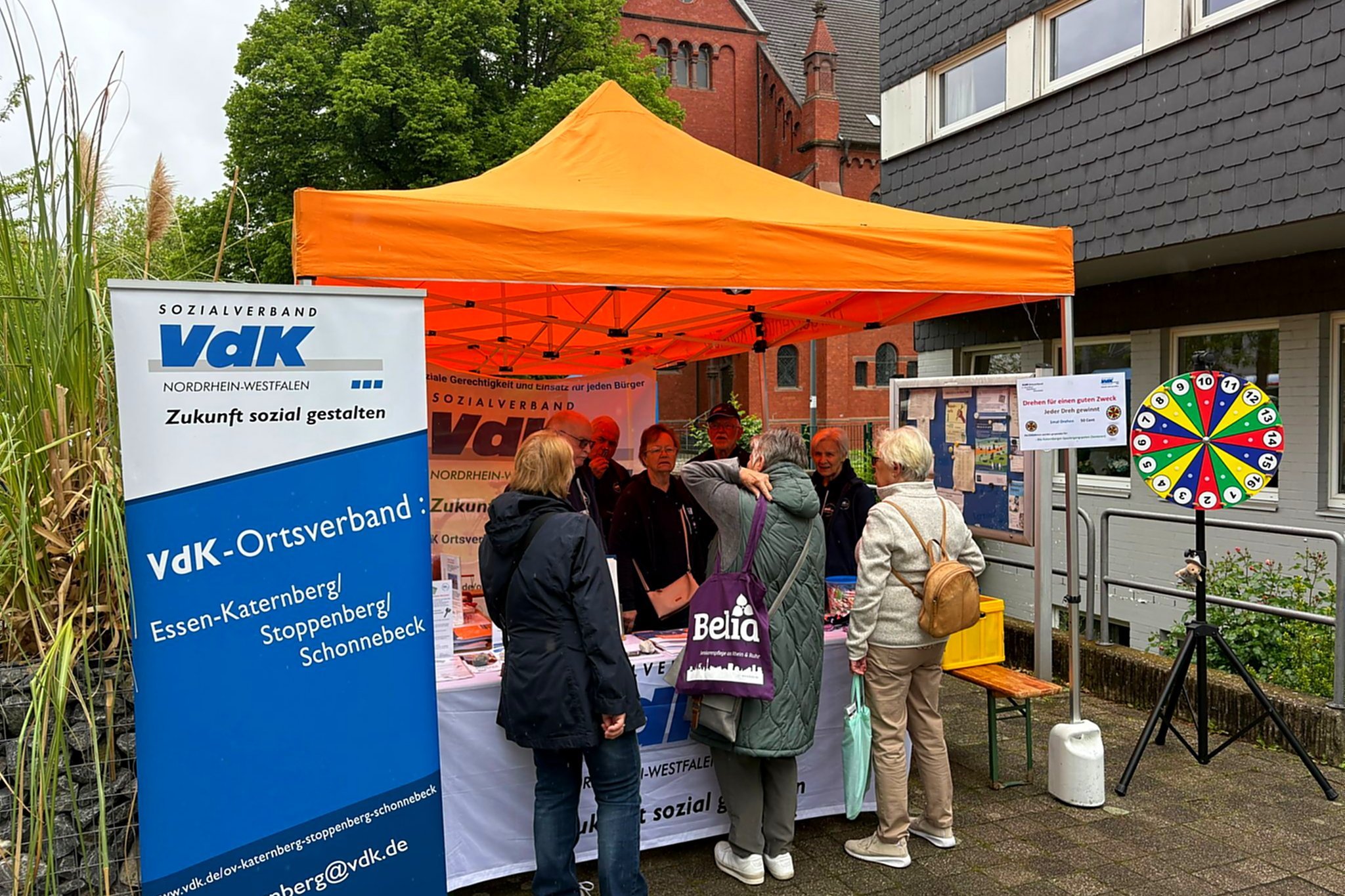 Drei Menschen stehen an einem VdK-Infostand mit orangem Pavillon. Rechts ein blau-weißes Roll-Up mit VdK-Logo, links ein buntes Glücksrad.
