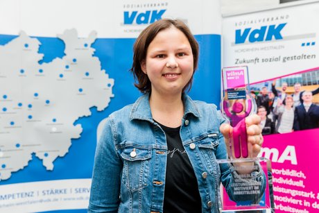 Preisträgerin Tabea Bigge hält den vilmaA-Pokal in die Höhe. Im Hintergrund Roll-Ups mit VdK-NRW-Logo und vilmA-Logo.