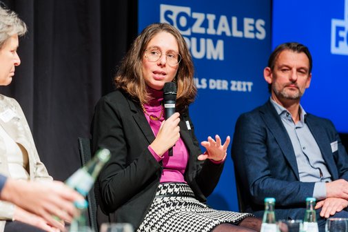 Eine Frau mit Brille und mittellangen braunen Haaren sitzt auf einer Bühne und spricht in ein Mikrofon.