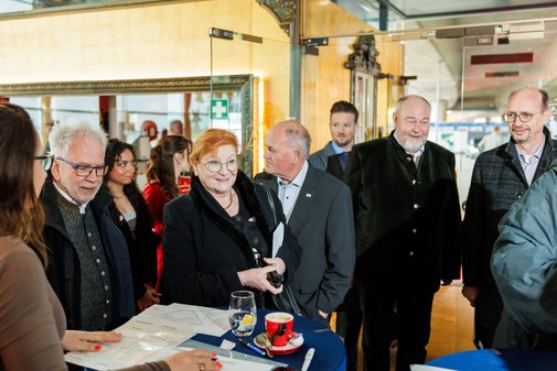 sieben Personen warten an einem Stehtisch auf ihre Anmeldung. Auf einem Stehtisch liegen viele Zettel.