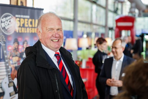 Herbert Hoffacker vom VdK M&ouml;nchengladbach l&auml;chelt in die Kamera. Hinter ihm unterhalten sich G&auml;ste der Veranstaltung.