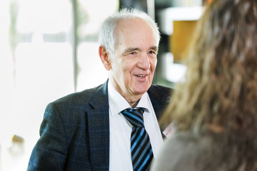 Ein Mann mit grauen Haaren spricht zu einer Frau mit langen Haaren. Der Mann tr&auml;gt ein Sakko mit einem wei&szlig;em Hemd und Krawatte.