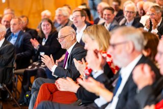 Menschen sitzen in den Stuhlreihen des Saals und applaudieren. 