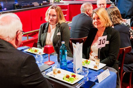 Zwei Frauen mit mittellangen blonden Haaren und ein älterer Herr mit weißen Haaren sitzen gemeinsam an einem Tisch bei einer Salatvorspeise. Im Hintergrund sind weitere Menschen an Tischen zu erkennen. 