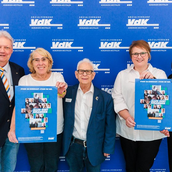 Zwei Männer und drei Frauen stehen nebeneinander vor einer blauen Wand mit weißen VdK-Logos. Zwei der Frauen halten ein Plakat der aktuellen Ehrenamtskampagne mit dem Slogan "Alles geht zusammen".