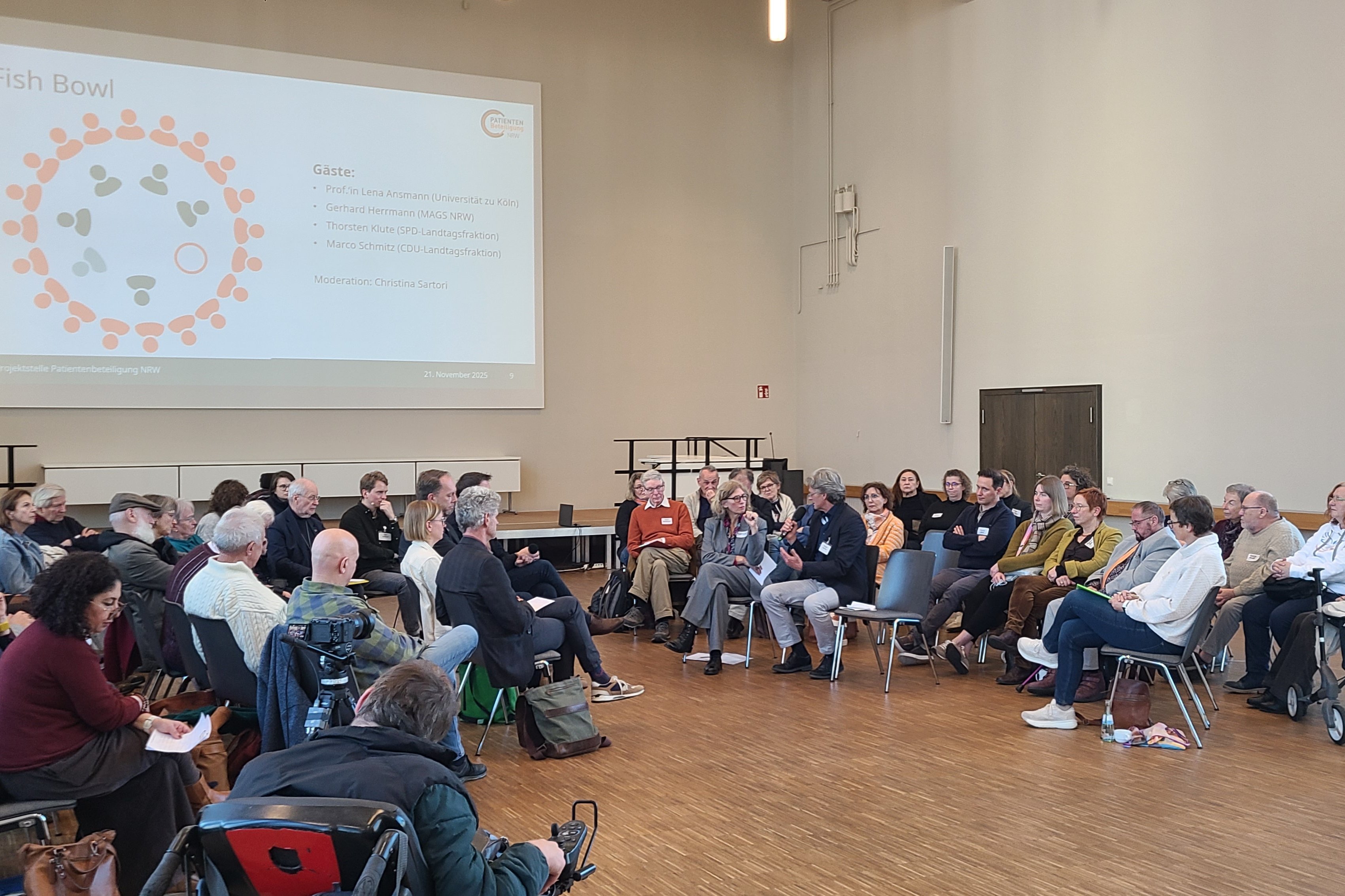 Viele Menschen sitzen in einem Saal. An der Wand ist eine Pr&auml;sentation eingeblendet. 