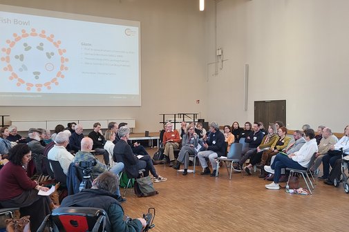 Viele Menschen sitzen in einem Saal. An der Wand ist eine Pr&auml;sentation eingeblendet. 