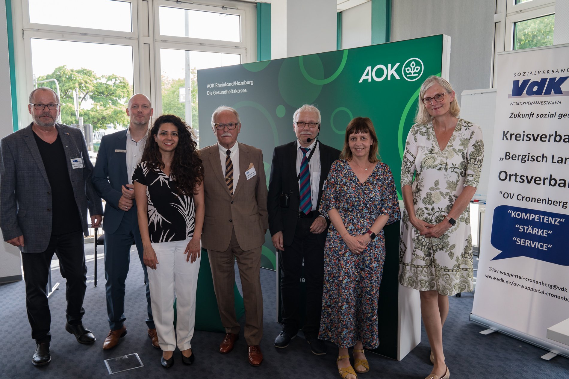 Gruppenfoto mit dem OV-Vorsitzenden und Referentinnen und Referenten des Abends vor einem gr&uuml;nen Banner der AOK und einem Roll-Up mit VdK-Logo.