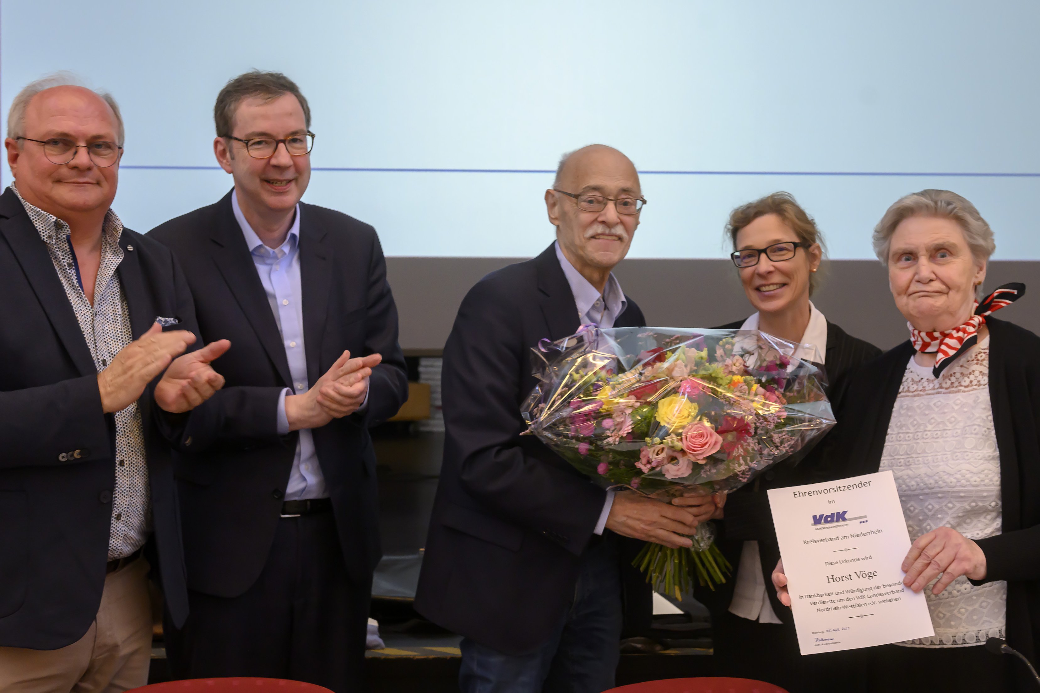 Drei Männer und zwei Frauen stehen nebeneinander. Der Mann in der Mitte bekommt von der Frau neben ihm einen Blumenstrauß überreicht. Die andere hält eine Ehrenurkunde in der Hand. Die beiden Männer links im Bild applaudieren.
