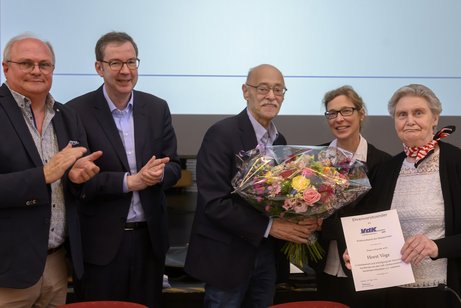 Drei Männer und zwei Frauen stehen nebeneinander. Der Mann in der Mitte bekommt von der Frau neben ihm einen Blumenstrauß überreicht. Die andere hält eine Ehrenurkunde in der Hand. Die beiden Männer links im Bild applaudieren.