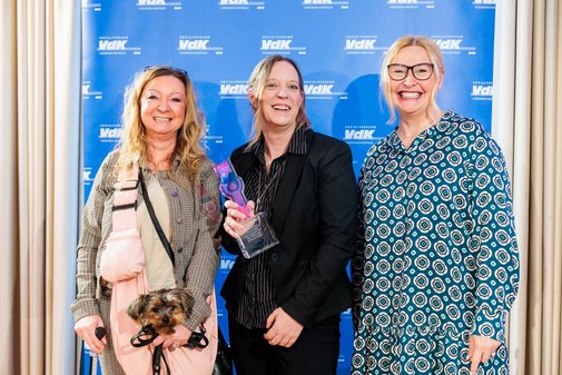 Eine vilmA-Presiträgerin und zwei Frauen rechts und links neben ihr vor einer blauen VdK-Wand. Eine der Frauen hat einen kleinen Yorkshire-Terrier auf dem Arm. Die Gewinnerin hält den vilmA-Pokal.