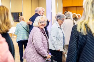 Eine Frau mit Rollator lächelt in die Kamera. Um sie herum laufen weitere Menschen durch das Foyer der Stadthalle Neuss.