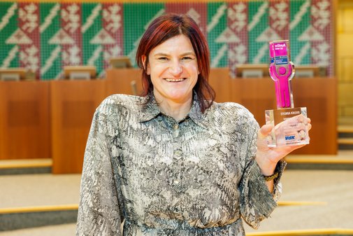 Eine Frau mit dunkelroten, mittellangen Haaren und gebl&uuml;mtem Kleid h&auml;lt l&auml;chelnd den vilmA-Preis in die H&ouml;he. 