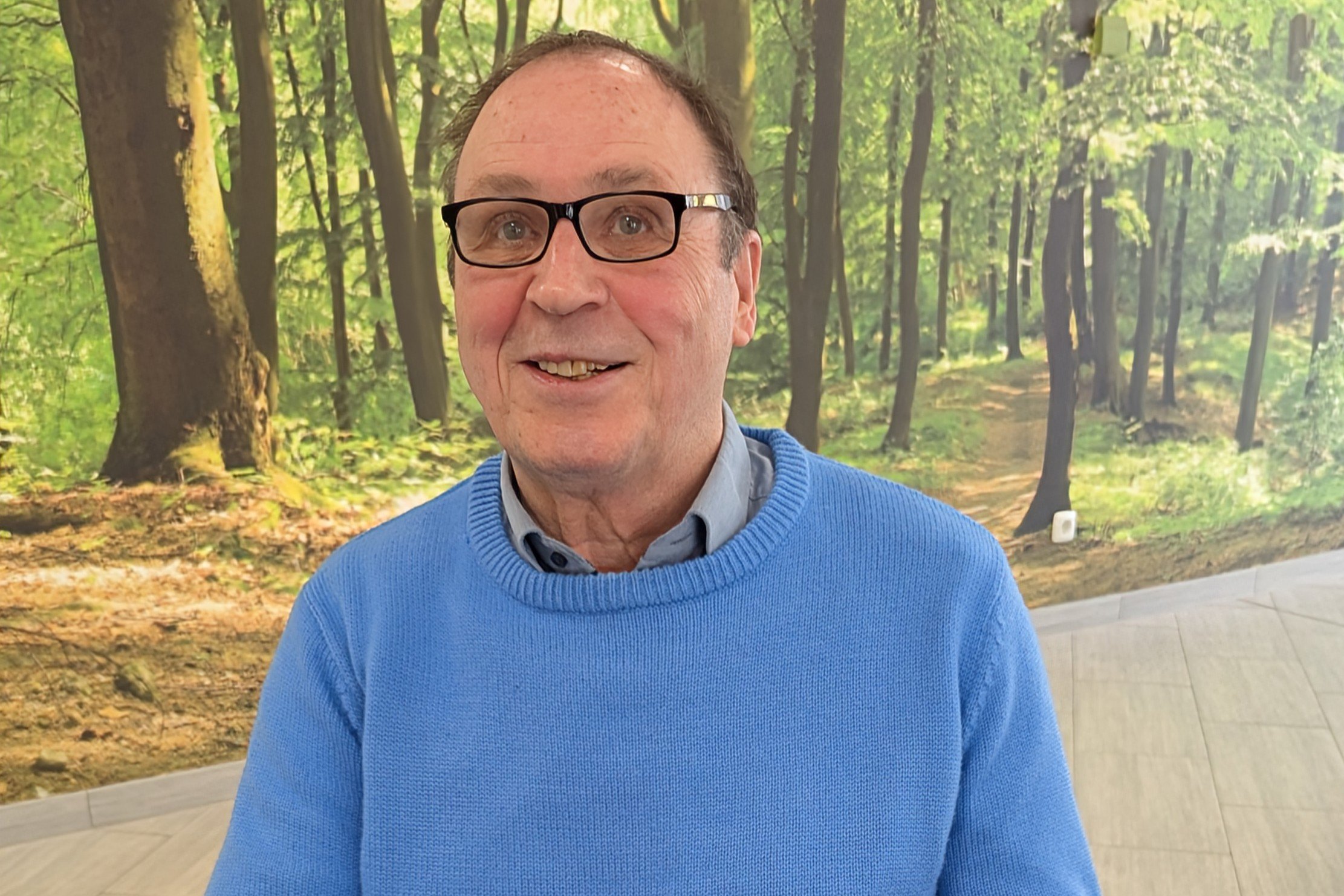 Ein älterer Mann mit Brille vor einer Fototapete, die einen lichtdurchfluteten Wald darstellt.