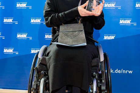 Die Preisträgerin Dana Kreuels sitzt im Rollstuhl vor einer blauen Wand mit VdK-Logos und hält lächelnd den vilmA-Pokal in die Höhe.