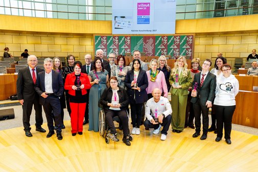 Eine große Gruppe Menschen, Männer und Frauen unterschiedlichen Alters, mit und ohne sichtbare Behinderung hat sich im Plenarsaal unter einer Leinwand mit vilmA-Logo versammelt.