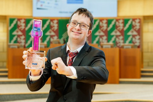 Ein junger Mann mit Brille und Kurzhaarfrisur h&auml;lt in der einen Hand den vilmA-Preis, die andere hat er zur Siegerfaust geballt und reckt sie Richtung Kamera.