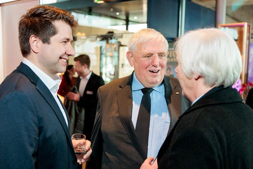 Zwei M&auml;nner mit Sakko unterhalten sich mit einer &auml;lteren Frau mit wei&szlig;en Haaren.