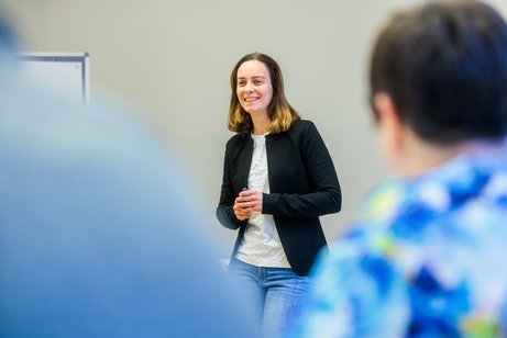 Eine Frau mit mittellangen, braunen Haaren in weißem Shirt, Jeans und schwarzem Sakko steht lächelnd vor Menschen, die von hinten fotografiert nur am Bildrand zu erkennen sind.
