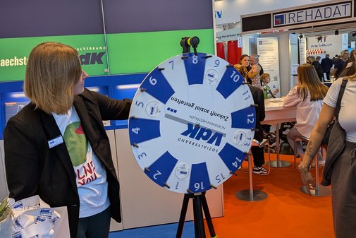 VdK-Referentin Vanessa Rengers-Patz steht neben dem Glücksrad mit VdK-Logo am Messestand.