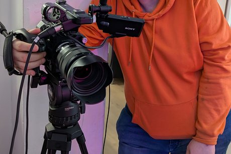 Ein Mann mit orangem Hoodie, Brille und großen Kopfhörern auf dem Kopf steht neben einer großen Stativ-Kamera.