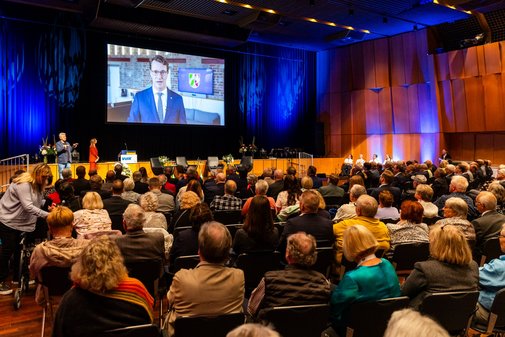 Menschen sitzen in einem Saal und schauen ein Video, dass per Beamer auf eine große Leinwand geworfen wird.