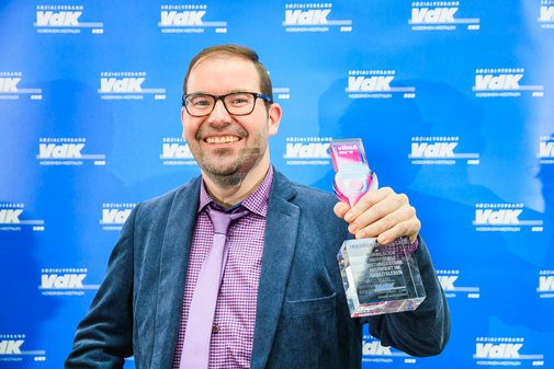 Ein lächelnder vilmA-Preisträger steht mit Pokal in der Hand vor einer blauen Wand mit weißen VdK-Logos. 